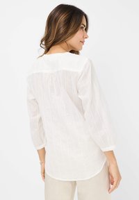 Blusa bianca a maniche lunghe con delicate strisce verticali, scollatura rotonda e maniche arricciate. Tessuto leggero con una vestibilità ampia e piega sul retro.