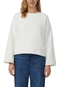 Biały, teksturowany sweter z okrągłym dekoltem, szerokimi rękawami i krótkim dołem, zestawiony z niebieskimi dżinsami. Ogólnie luźny krój.