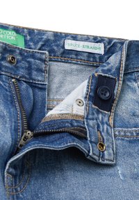 Jeans de denim con cremallera abierta y cierre de botones, mostrando las costuras internas y etiquetas de tejido que dicen "BRUCE-STRAIGHT" y "United Colors of Benetton."