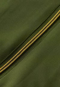 Tessuto verde oliva caratterizzato da una zip gialla prominente lungo la cucitura, con una superficie liscia e dettagli di cucitura precisi.