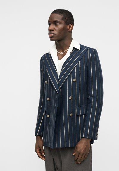Άνδρας ντυμένος με διπλόμπροκο σακάκι σκούρου μπλε με χρυσές κάθετες ρίγες, λευκό πουκάμισο, μαργαριταρένιο κολιέ και γκρι παντελόνι, στέκεται μπροστά σε λευκό φόντο.
