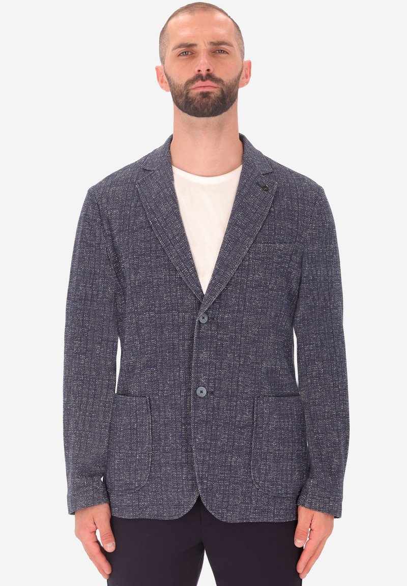 Uomo che indossa un blazer strutturato blu scuro su una camicia bianca e pantaloni scuri, in piedi contro uno sfondo bianco uniforme.