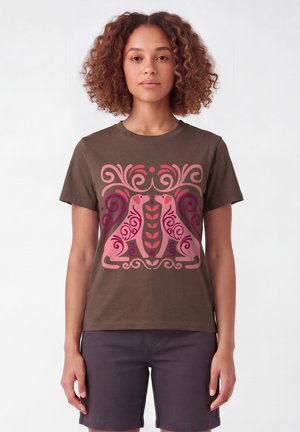 Vrouw draagt een bruin T-shirt met een roze en paarse symmetrische afbeelding van twee gestileerde zittende honden die elkaar tegenover elkaar aankijken.