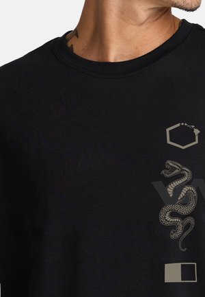 Nahaufnahme einer Person, die ein schwarzes Shirt mit einem Schlangengrafikmotiv, einer Sechseckform und einem kleinen Quadrat auf der rechten Vorderseite trägt.