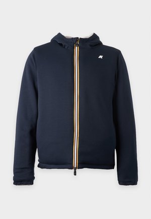 Mand iført en mørkeblå hættetrøje-jakke over en sort poloshirt, lyseblå vidde buksere og hvide sneakers, stående mod en enkel baggrund.