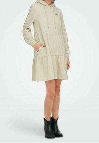 Vestido con capucha de color crema con diseño en capas. Cuenta con un bolsillo tipo canguro y texto bordado en verde. Hecho de tela suave.