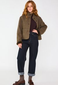 Giacca a scacchi marrone con alto colletto, jeans scuri, orlo risvoltato, mocassini marroni e calzini grigi; tessuto testurizzato e dettagli con bottoni visibili.