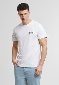Hvit bomull t-skjorte med rund hals, kortermet, og en svart "pray" tekstdesign på brystet. Kombinert med lyseblå jeans.