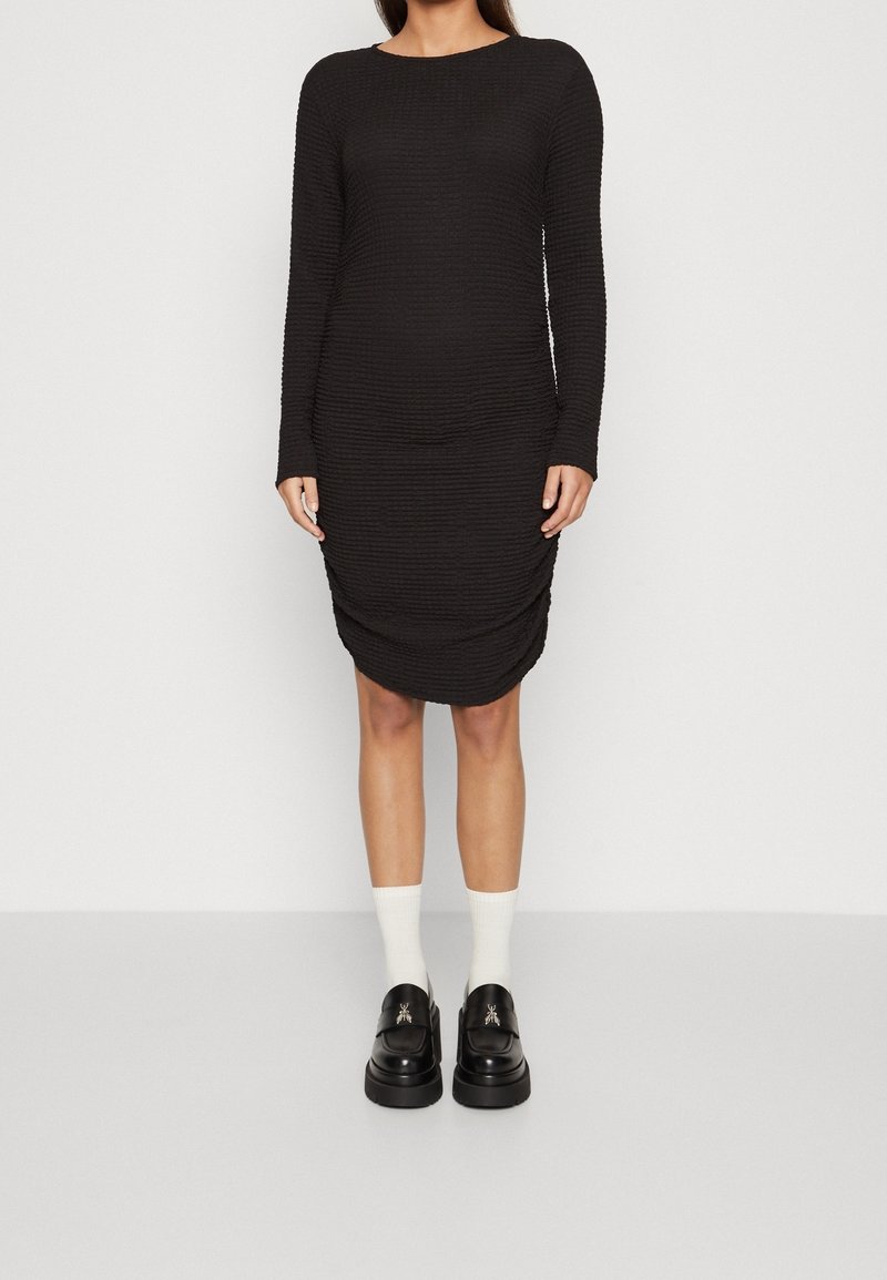Femme portant une robe noire texturée à manches longues, longueur genou, des chaussettes blanches et des mocassins noirs avec des détails argentés d'insecte, debout sur un sol clair.
