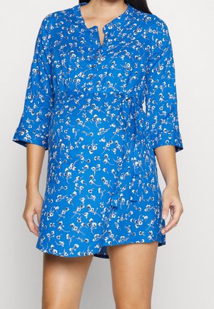 Vrouw die een blauwe bloemenjurk draagt met driekwartmouwen en een strik in de taille, staand tegen een effen lichte achtergrond.