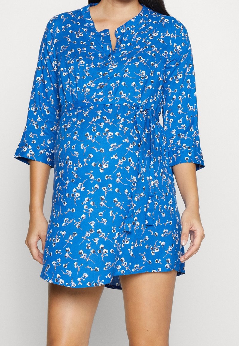 Mujer vestida con un vestido azul de flores, con mangas tres cuartos y un lazo en la cintura, de pie frente a un fondo claro y liso.