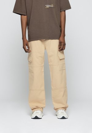 T-shirt oversized marron avec logo sur la poitrine, associé à un pantalon cargo beige avec poches latérales et coupe détendue ; sneakers blanches.