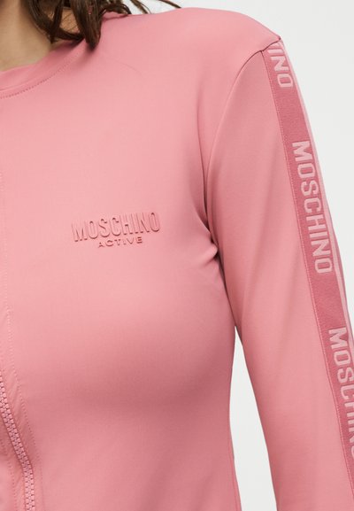 Κοντινή λήψη ενός ροζ αθλητικού μακρυμάνικου τοπ με το λογότυπο "Moschino Active" στο στήθος και τη σήμανση στη ρίγα του μανικιού.