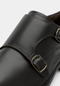 Sapata de couro preta com uma tira larga e fivela prateada, textura suave e costura subtil ao longo das bordas.