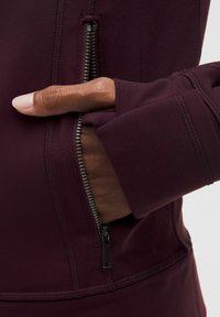 Burgundowa kurtka z zamkiem, z ręką w kieszeni, o miękkiej fakturze i bezszwowych mankietach dla komfortu i funkcjonalności.