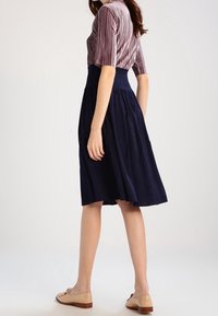 Woman wearing a mauve ribbed top tucked into a high-waisted navy pleated midi skirt with beige flat shoes standing sideways.