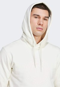 Weiße Kapuzenjacke aus weichem Stoff, mit einer Kordelzugkapuze und einem dezenten, eingeprägten Logo auf der Vorderseite. Glatte Textur, figurbetontes Design.