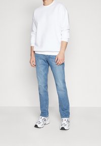 Man som bär en enkel vit sweatshirt, ljusblå jeans och vita sportskor, står mot en neutral bakgrund.