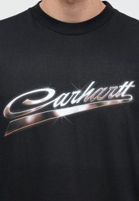 Czarny bawełniany t-shirt z metalicznym, srebrno-pomarańczowym logo „Carhartt” z błyszczącym efektem na jednolitym tle.
