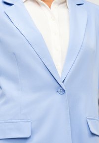 Ανοιχτό μπλε σακάκι με ένα κουμπί πάνω από λευκό ριμπ μπλουζάκι με κουμπιά, εμφανίζεται από το στήθος έως το ύψος της μέσης.