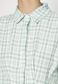 Camisa xadrez verde e branca com colarinho, apresentando uma frente abotoada e uma textura de tecido macio. Exibe padrões de grelha e plaid.