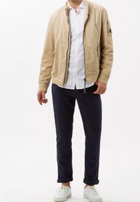 Beige lätt bomberjacka med dragkedja framtill, sidofickor, ribbade ärmslut och ett märke på vänster arm, kombinerat med marinblå chinos och vita sneakers.