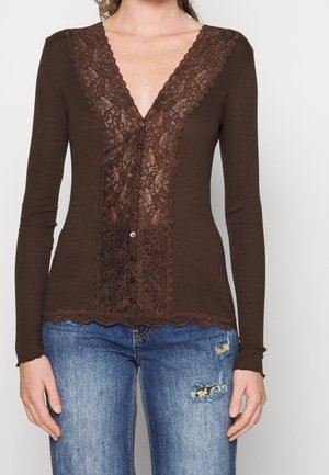 Mujer con camiseta marrón de manga larga con panel de encaje y botones delanteros, combinada con vaqueros azules desgastados.