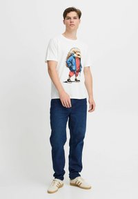 Hvid grafisk t-shirt med en pindsvin i en blå jakke og røde bukser. Båret med mørkeblå jeans og hvide sneakers med gyldne accenter.