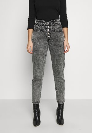 Person trägt schwarze High-Waist Acid-Wash-Jeans mit silbernen Knöpfen und schwarze Stiefeletten und steht vor einem schlichten Hintergrund.