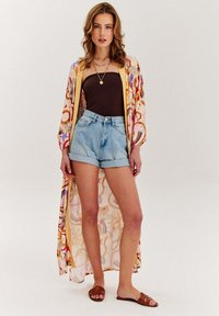 Dlouhý, vzorovaný kimono v pastelových barvách přes hnědý top a džínové šortky. Měkká tkanina, otevřený střih a volný design. Hnědé sandály dotvářejí outfit.