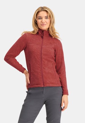 Frau trägt eine bordeauxrote Fleecejacke mit Reißverschluss und graue Hose, steht mit einer Hand an der Hüfte vor einem einfarbigen Hintergrund.