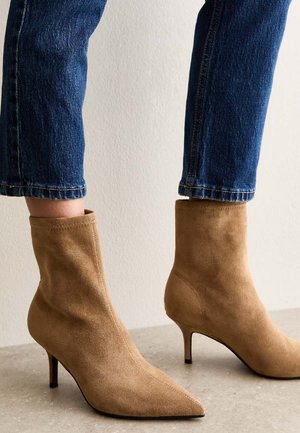 Botines de ante color canela con punta afilada y tacón stiletto. Los botines tienen una textura suave y un diseño ajustado, combinados con vaqueros azules recortados.