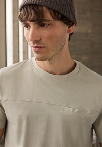 Jasnobeżowa koszulka wykonana z miękkiego materiału, z okrągłym dekoltem, krótkimi rękawami oraz małą kieszonką na piersi z subtelnym poziomym szwem.