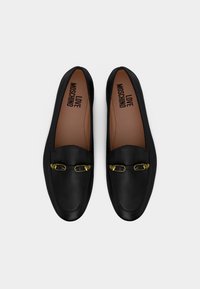 Μαύρα δερμάτινα μοκασίνια με χρυσές μεταλλικές λεπτομέρειες σε σχήμα καρδιάς στην κορυφή, ταμπά εσωτερικά πέλματα με τη μάρκα "LOVE MOSCHINO", εμφανίζονται από πάνω.