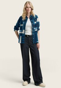 Camicia a maniche lunghe blu e bianca a fantasia con maniche arrotolate, indossata sopra un top bianco e jeans a gamba larga scuri con cintura.