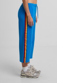 Modré tepláky s bočním pruhem v červené, žluté a oranžové barvě. Mají elastický pas a volný střih, doplněné šedými teniskami.