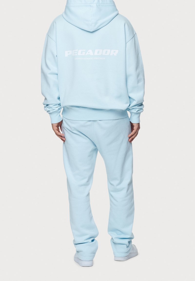 Personne portant un sweat à capuche bleu clair et un pantalon de survêtement assorti, debout dos tourné, texte blanc "PEGADOR" au dos du sweat à capuche.