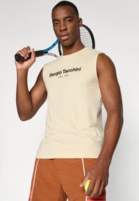 Beige ermeløs bomull T-skjorte med svart "Sergio Tacchini EST. 1966" logo på fronten, iført brune shorts, holder en tenniskølle og ball.