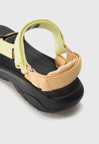 Zwarte zool met een gestructureerd patroon, voorzien van gele en perzikkleurige verstelbare banden. Zachte padding op de hak voor comfort. Lichte constructie.