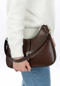 Brown leather shoulder bag with a curved shape, featuring a gold-tone hardware accent and a short strap, held by a person in denim.
