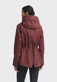 Veste extérieure bordeaux avec taille ajustable, grande capuche et manches longues. Texture lisse, coupe mi-longue et détail à l'arrière avec une fente.