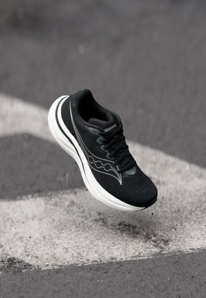 Melns Saucony skriešanas apavu ar baltu zolīti gaisā virs pelēka asfalta ar baltu ceļa marķējumu.
