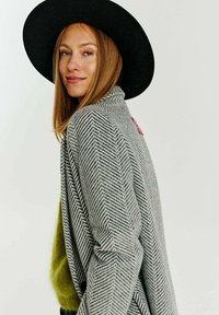 Šedý kabát s herringbone vzorem a volným střihem, nošený přes živě zelený svetr. Černý klobouk s širokým krempou dokončuje vzhled.