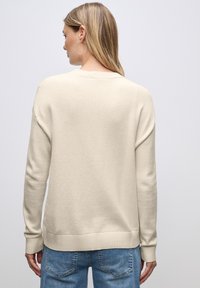 Cremfarbener Pullover mit geripptem Saum und Bündchen, der ein einfaches Design und eine glatte Textur aufweist. Über einer hellblauen Jeans getragen.