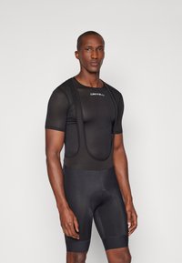 Melna velosipēdu jakas ar acu struktūru un plecu siksnām, apvienota ar melnām velosipēdu biksēm, kurām ir polsterēta sēdvieta.