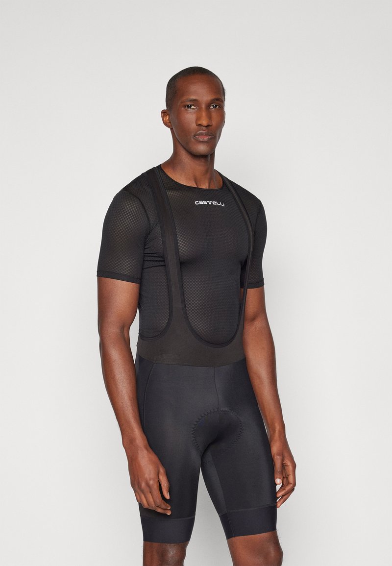 Melna velosipēdu jakas ar acu struktūru un plecu siksnām, apvienota ar melnām velosipēdu biksēm, kurām ir polsterēta sēdvieta.