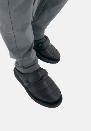 Persoon met zwarte gevoerde instapschoenen met verstelbare bandjes en donkergrijze broek tegen een lichte achtergrond.