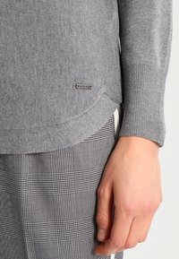 Grå strikket sweater med ribbede manchetter og en subtil logo-detail. Kombineret med sort- og hvidternet stof, hvilket viser teksturkontrast.