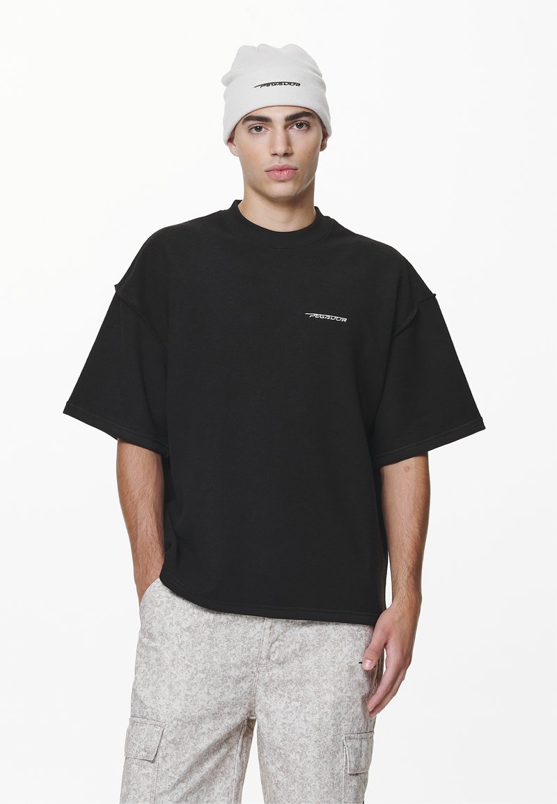 Maglietta nera oversize a maniche corte con un piccolo logo bianco. Abbinala a pantaloni cargo chiari beige con motivo e a un berretto bianco.