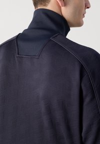 Tmavomodrý sweatshirt s vysokým golierom, ktorý má textúrovaný geometrický vzor, ribované tkaniny a detaily na švom na pleciach.